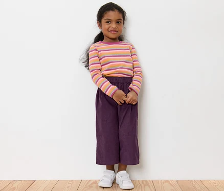 Ein Mädchen steht und trägt eine Kinder-Culotte, ein gestreiftes T-Shirt und weiße Schuhe.