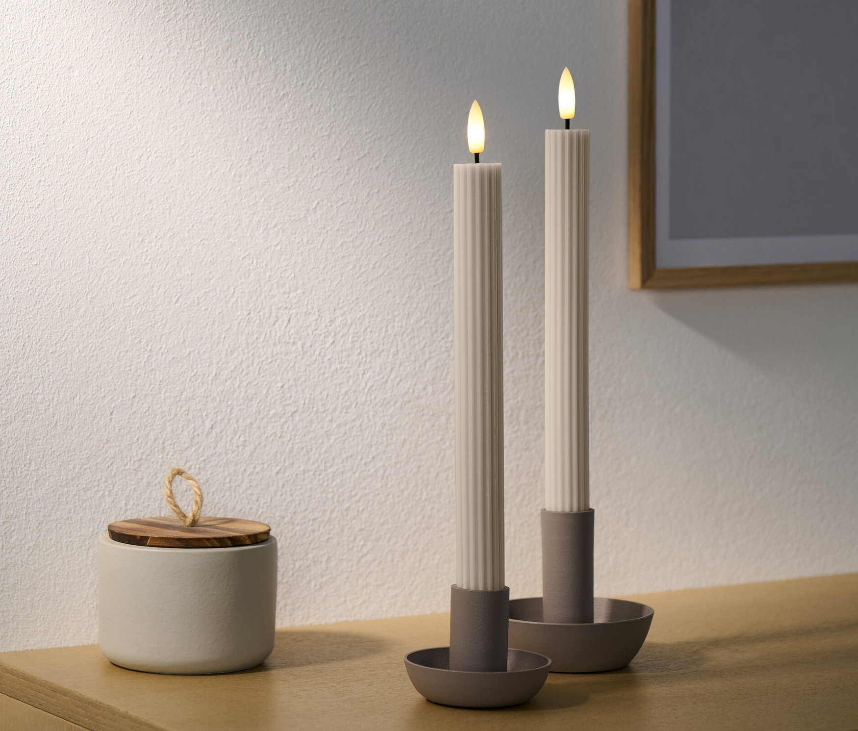Zwei gerillte LED-Echtwachs-Stabkerzen auf Ständern stehen auf einem Schrank neben einer Dose.