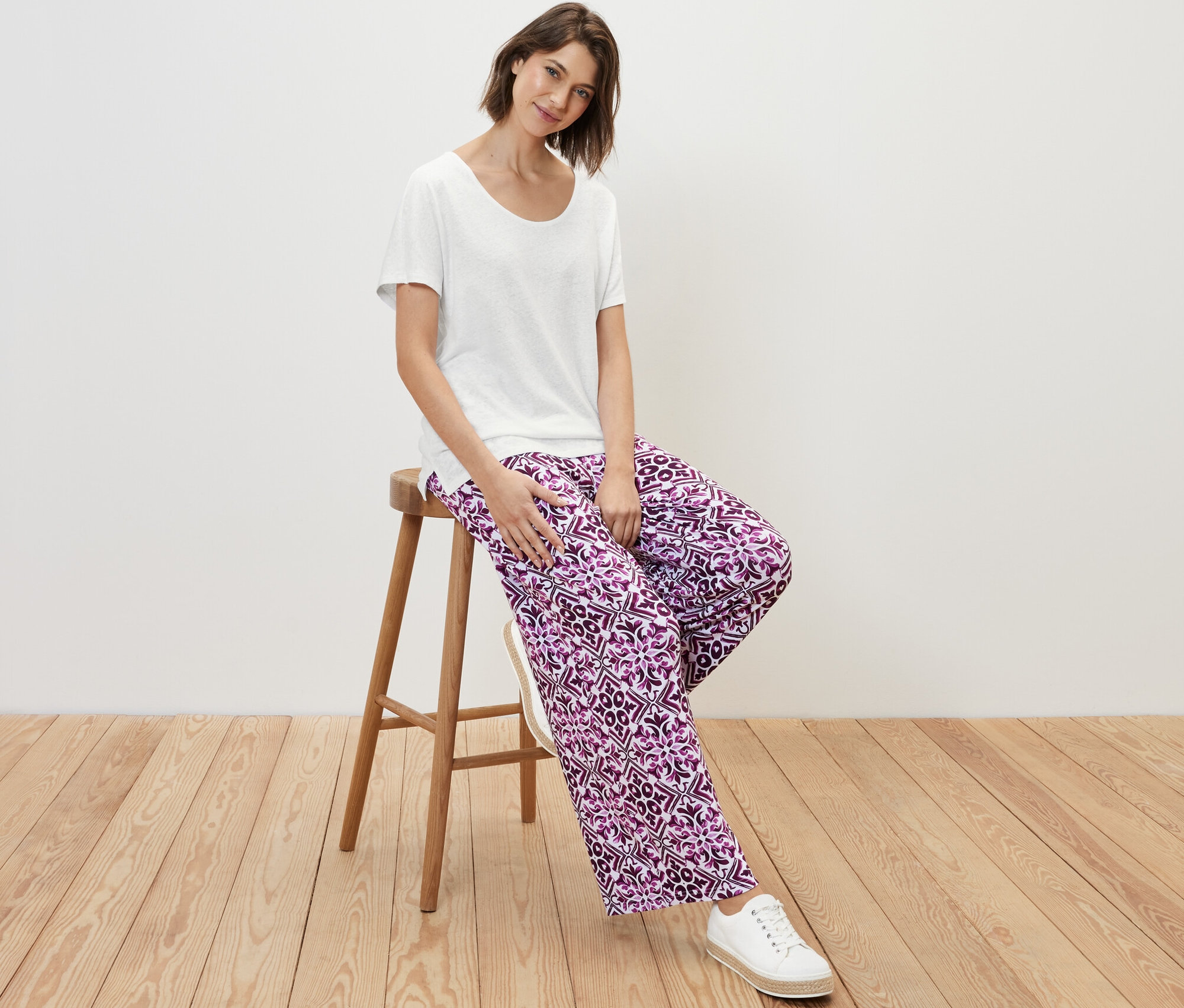 Frau sitzt auf einem Hocker und trägt ein weißes Shirt aus Leinenmix und gemusterte Hosen.