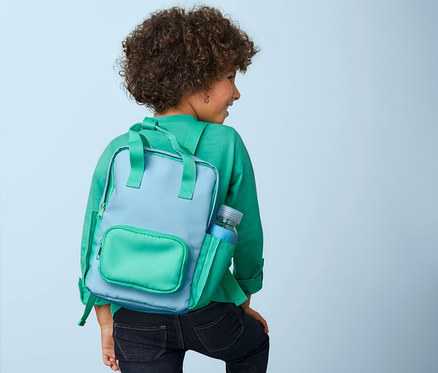 Ein Junge mit lockigem Haar steigt Stufen hinauf und trägt einen blau-grünen Outdoor-Rucksack, mini.