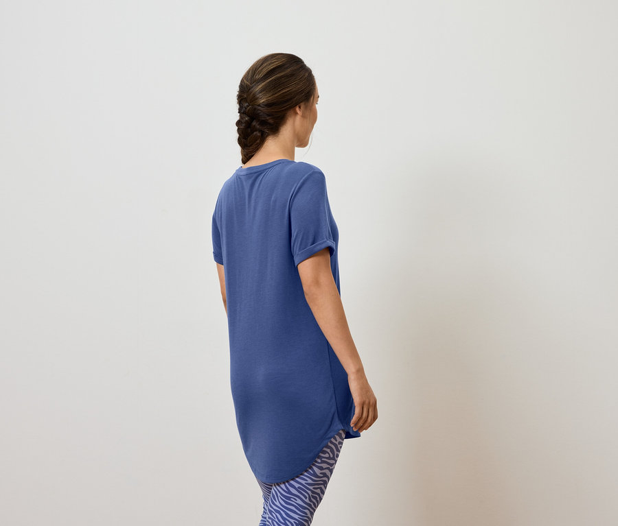 Frau mit Zopf trägt ein blaues T-Shirt und gemusterte Leggings, von hinten gesehen.