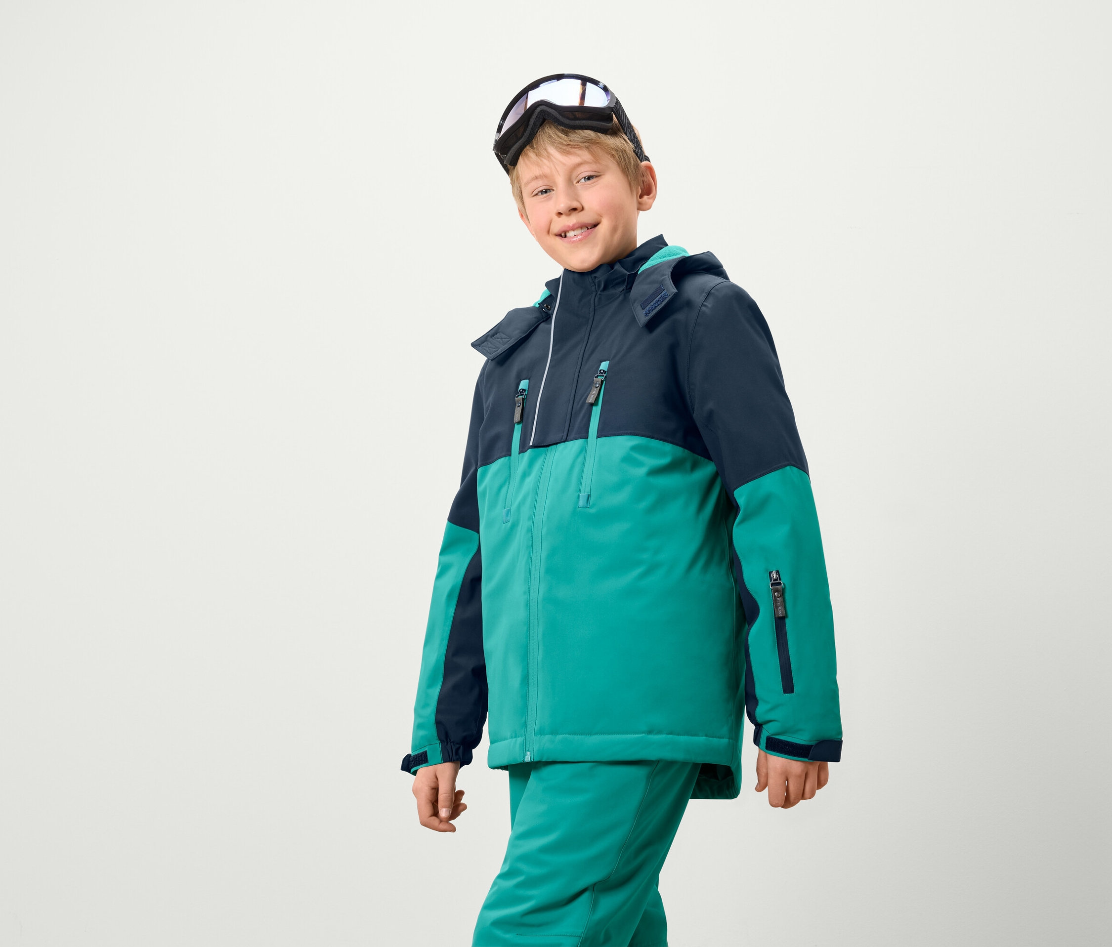 Ein Junge trägt eine Skijacke und -hose, mit einer Skibrille auf dem Kopf.