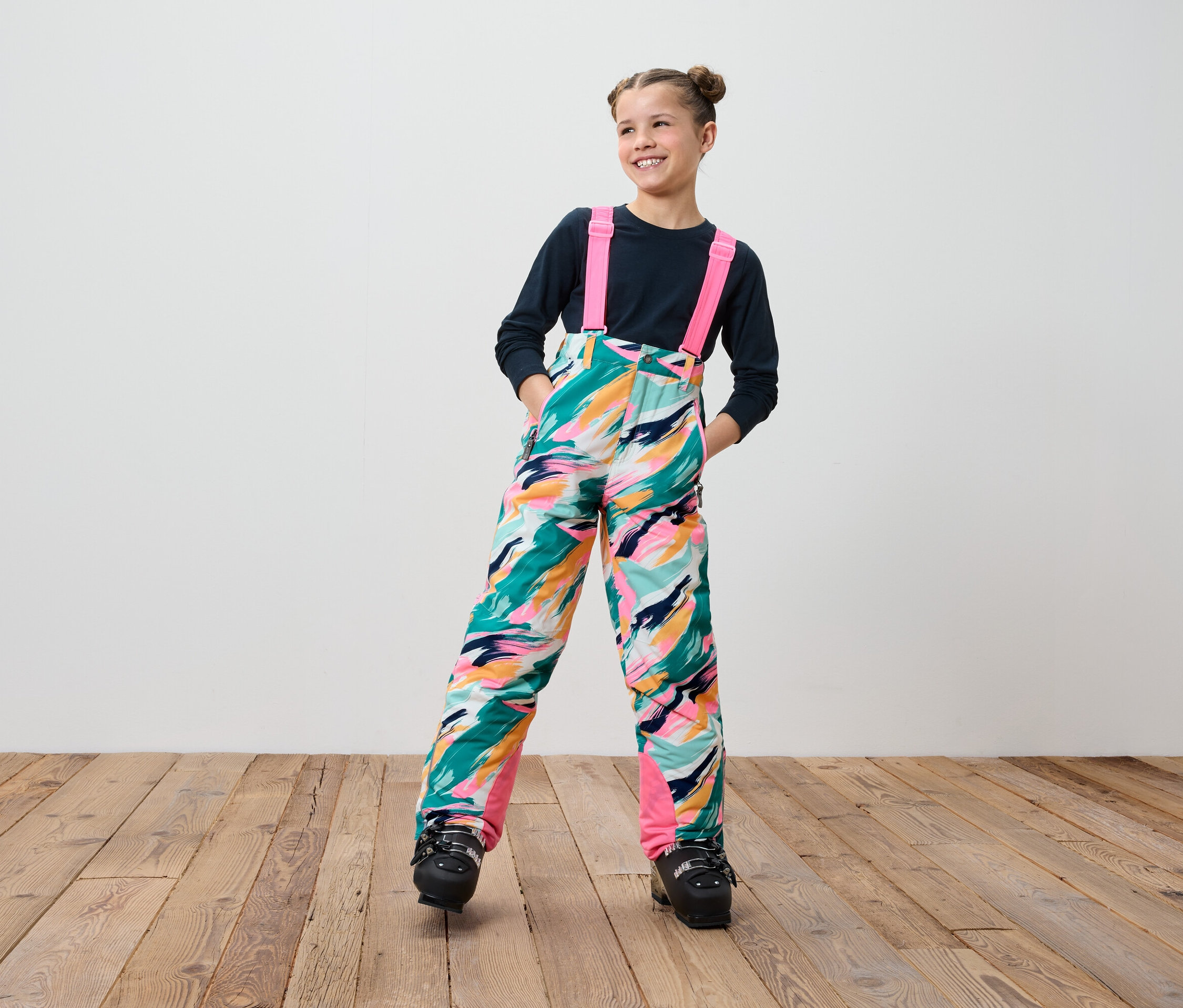 Lächelndes junges Mädchen in farbenfroher Skihose mit Hosenträgern und schwarzen Skischuhen steht auf einem Holzboden.