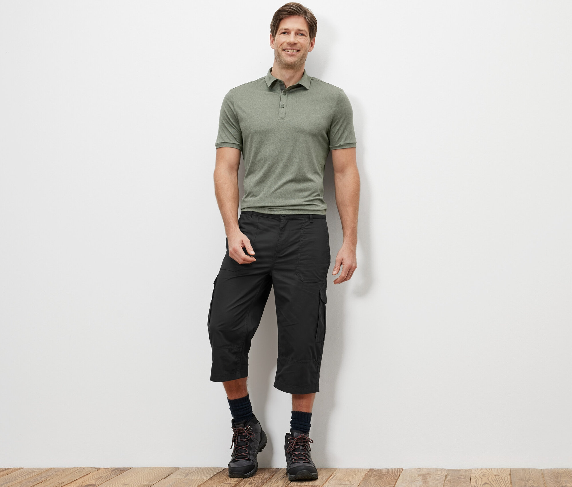 Ein Mann steht und trägt ein grünes Poloshirt und eine anthrazitfarbene Cargo-Bermuda in 3/4-Länge.