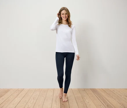 Eine Frau geht barfuß über einen Holzboden und trägt ein weißes Langarmshirt und dunkelblaue Leggings.