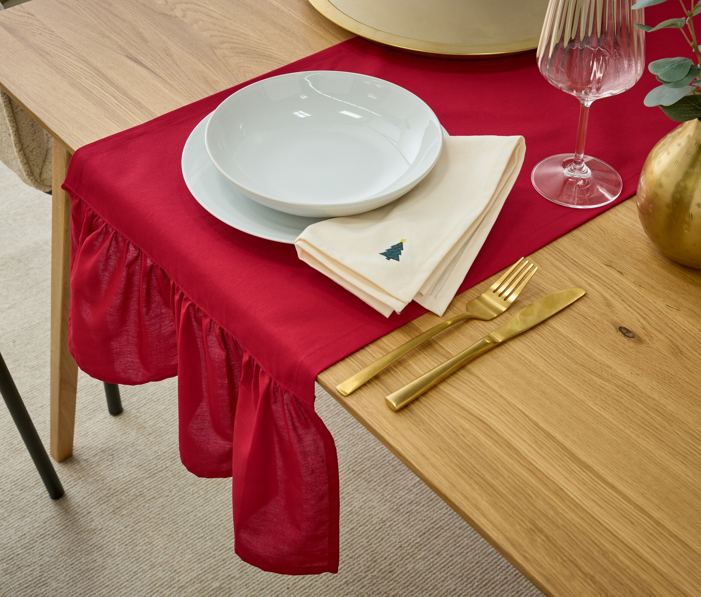 Gedeckter Tisch mit roter Tischdecke, weißem Geschirr, beiger Serviette mit Weihnachtsbaum-Motiv, goldenem Besteck, Weinglas und goldener Vase mit Eukalyptus.