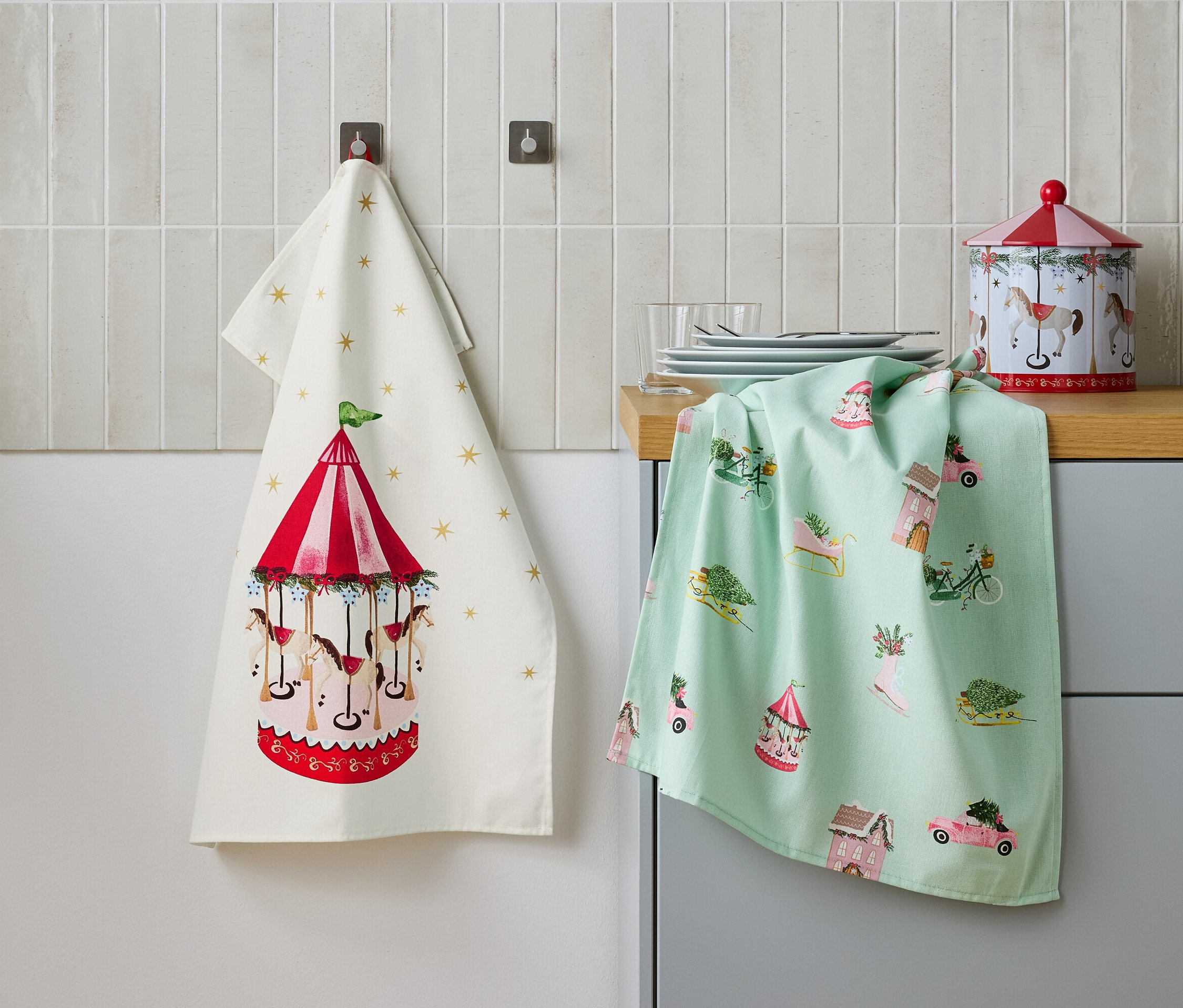 Zwei Geschirrtücher hängen nebeneinander an einer Wand, eines mit einem Karussellmotiv und das andere mit festlichen Motiven, sowie eine Blechdose auf einem Tisch.