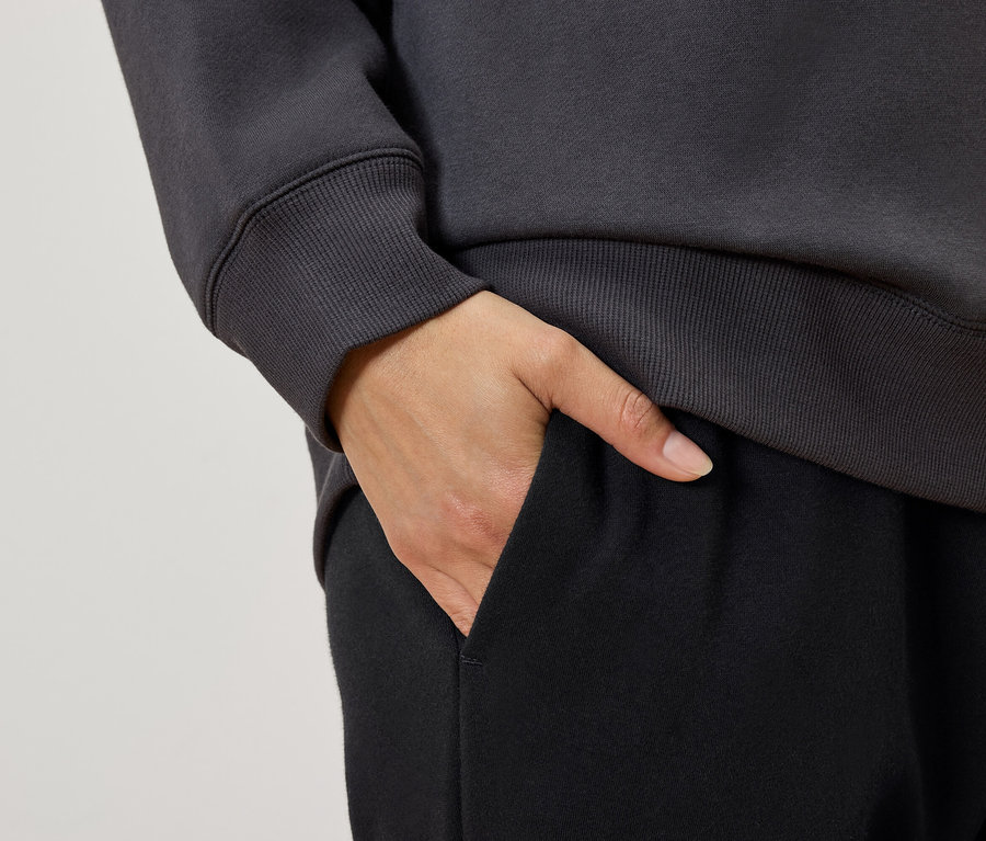 Detailaufnahme einer Frau mit der Hand in der Tasche einer schwarzen Trainingshose, dazu ein graues Sweatshirt.