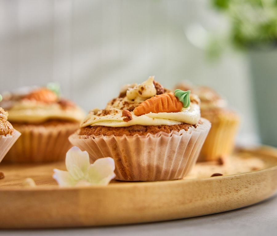 Vier Karottenmuffins mit Guss und Marzipanmöhren auf einem Holztablett.