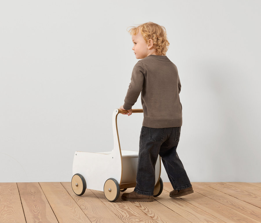 Kleiner Junge schiebt einen weißen Wagen mit braunen Rädern auf einem Holzboden.