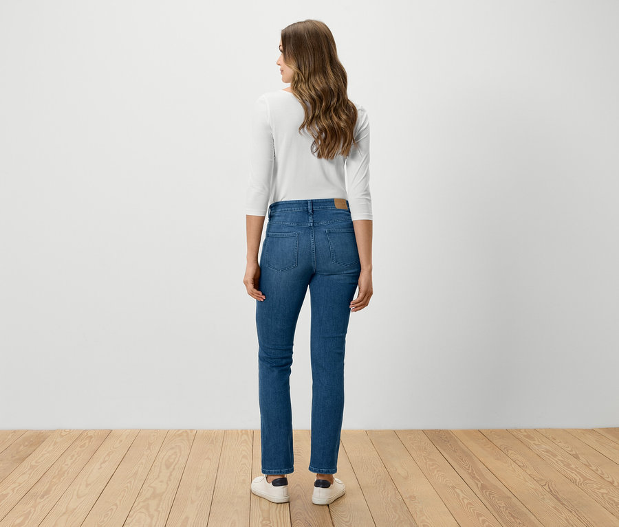 Eine Frau steht mit dem Rücken zur Kamera und trägt blaue Jeans, ein weißes Hemd und weiße Schuhe.
