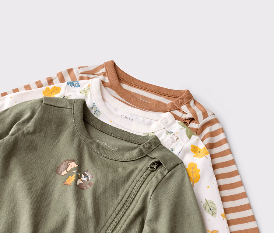 Drei Baby-Schlafanzüge liegen übereinander, einer olivgrün mit Tiermotiv, einer weiß mit Blättern und einer gestreift.