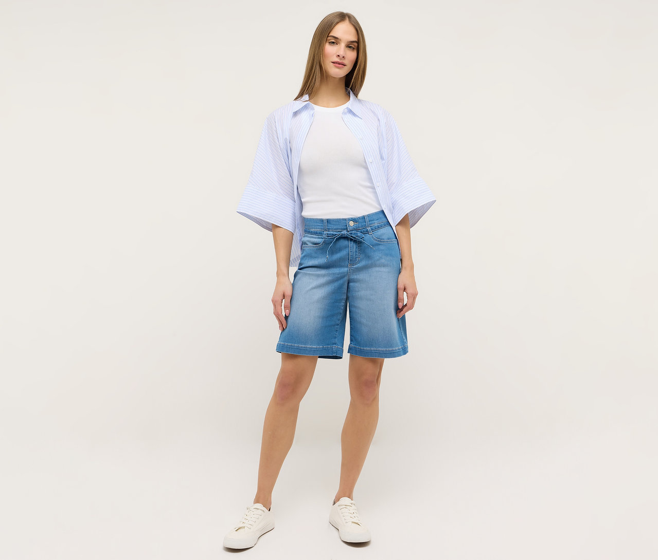 Frau in Jeans »Lia City« Shorts von Angels in Hellblau, weißem Shirt, blau gestreifter Bluse und weißen Turnschuhen.