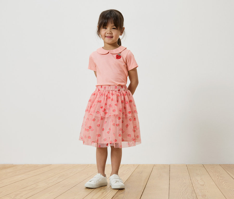 Ein junges Mädchen steht vor einer weißen Wand und trägt ein rosa T-Shirt mit Erdbeermotiv, einen rosa Tüllrock mit Erdbeeren und weiße Turnschuhe.
