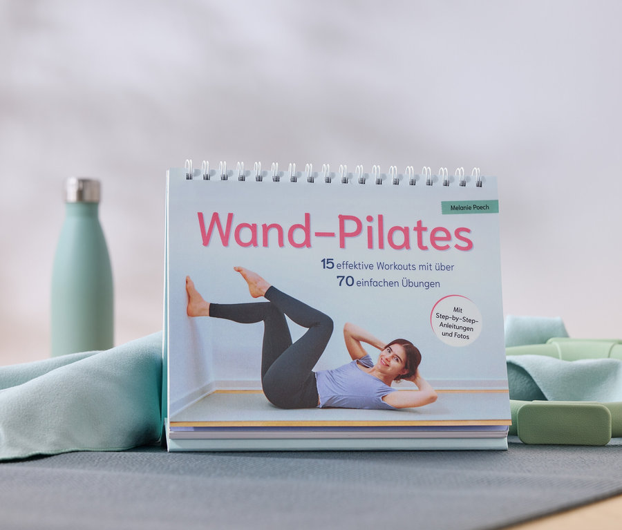 Ein Wand-Pilates Buch mit einer Frau, die eine Übung macht, daneben eine Trinkflasche, ein Handtuch, ein Pilates-Ring und ein Block.