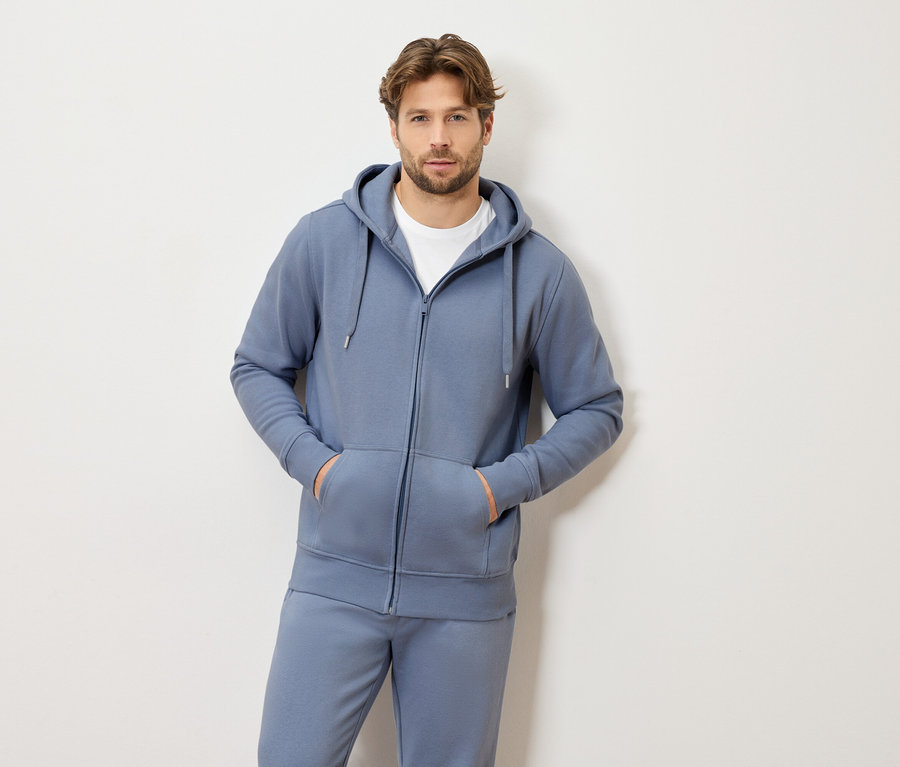 Mann mit hellbraunem Haar, der einen blauen Trainingsanzug mit Kapuze und Reißverschluss und ein weißes T-Shirt trägt und mit beiden Händen in den Taschen steht.