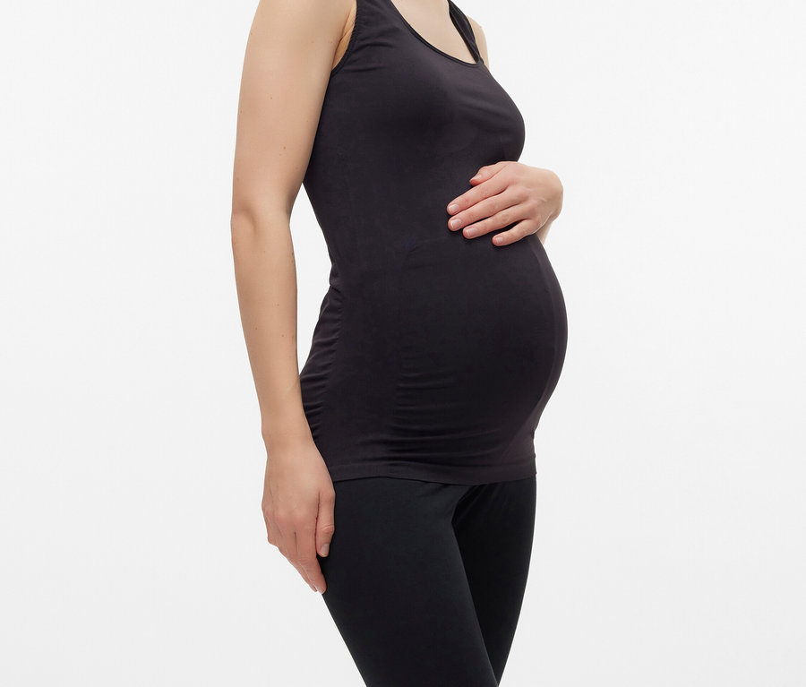 Schwangere Frau mit schwarzem Tanktop und Leggings posiert vor einem weißen Hintergrund.