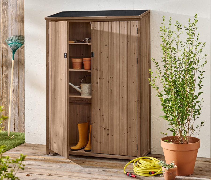 Ein offener Holzschrank steht draußen mit Gartengeräten, Töpfen, Gummistiefeln und einer Gießkanne im Inneren.