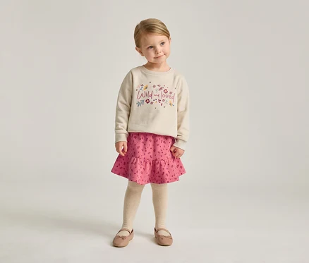 Ein kleines Mädchen mit blonden Haaren trägt ein beiges Sweatshirt mit der Aufschrift "Wild and loved", einen rosa Rock und Ballerinaschuhe.