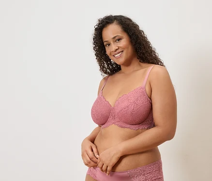 Frau mit lockigem Haar, die einen rosa Spitzen-BH und passende Unterwäsche vor einem schlichten Hintergrund trägt.