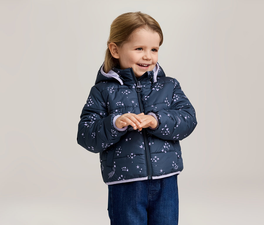 Lächelndes Mädchen mit blonder Frisur trägt eine marineblaue, gesteppte Jacke mit Kapuze und Sternenmuster und blaue Jeans.