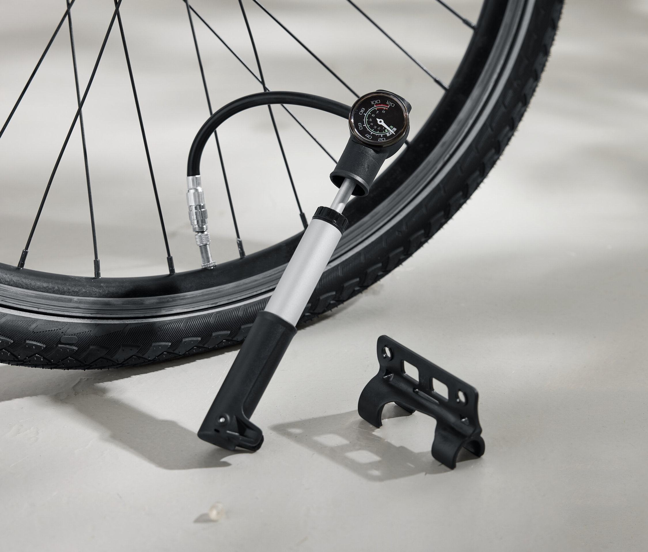 Ein Fahrradreifen wird mit einer silbernen Luftpumpe aufgepumpt, die über einen schwarzen Griff und einen analogen Manometer verfügt. Eine schwarze Halterung liegt daneben auf dem Boden.