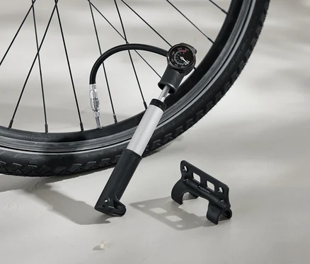 Ein Fahrradreifen wird mit einer silbernen Luftpumpe aufgepumpt, die über einen schwarzen Griff und einen analogen Manometer verfügt. Eine schwarze Halterung liegt daneben auf dem Boden.