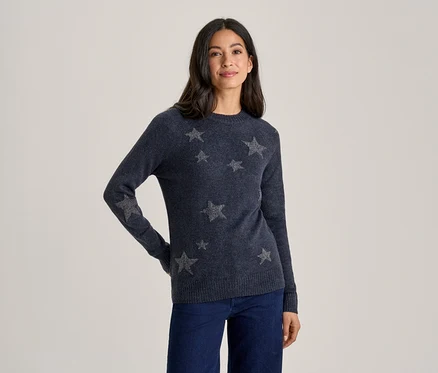 Eine Frau steht lächelnd mit den Händen in den Hüften und trägt einen dunkelblauen Pullover mit silbernen Sternen und eine dunkelblaue Jeans.