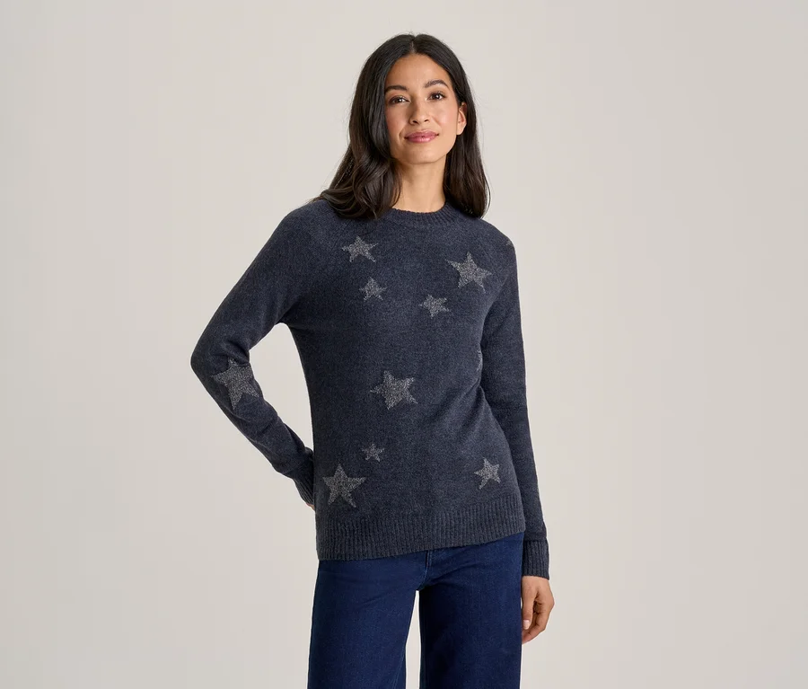 Eine Frau steht lächelnd mit den Händen in den Hüften und trägt einen dunkelblauen Pullover mit silbernen Sternen und eine dunkelblaue Jeans.