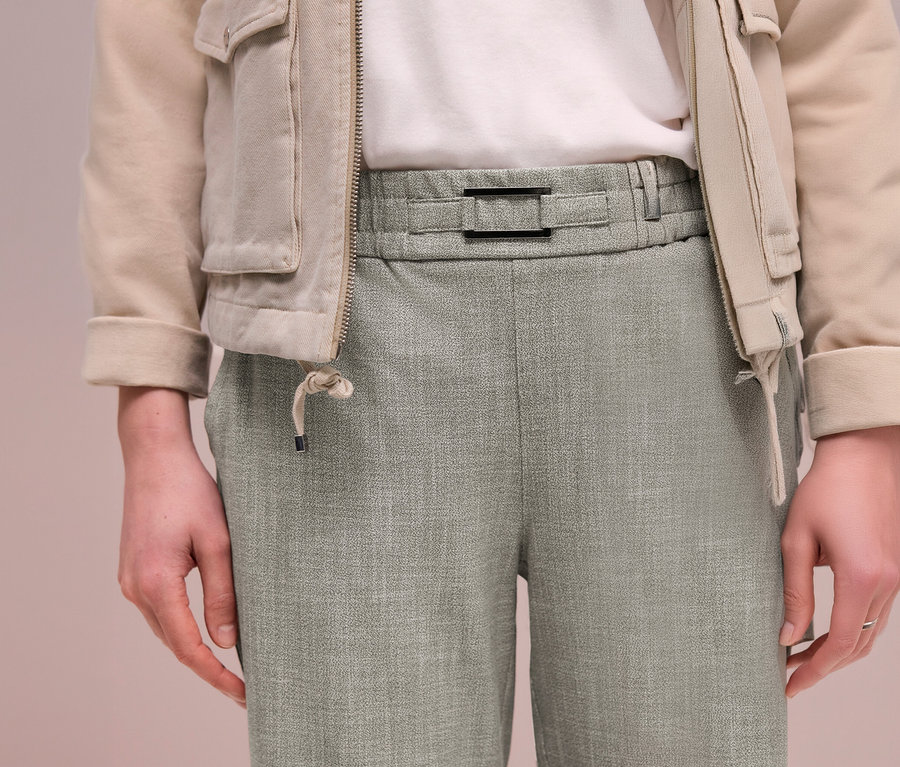 Person trägt eine beige Jacke über einem weißen T-Shirt und eine hellgrüne Street One Joggpants mit schwarzer Schnalle am Bund.