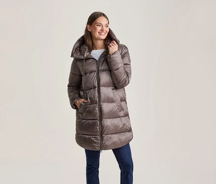 Lächelnde Frau in einer glänzenden braunen langen Steppjacke und blauen Jeans, eine Hand in der Tasche.