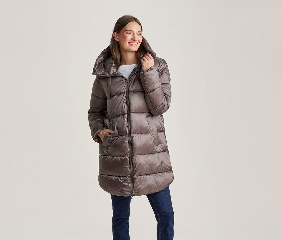 Lächelnde Frau in einer glänzenden braunen langen Steppjacke und blauen Jeans, eine Hand in der Tasche.