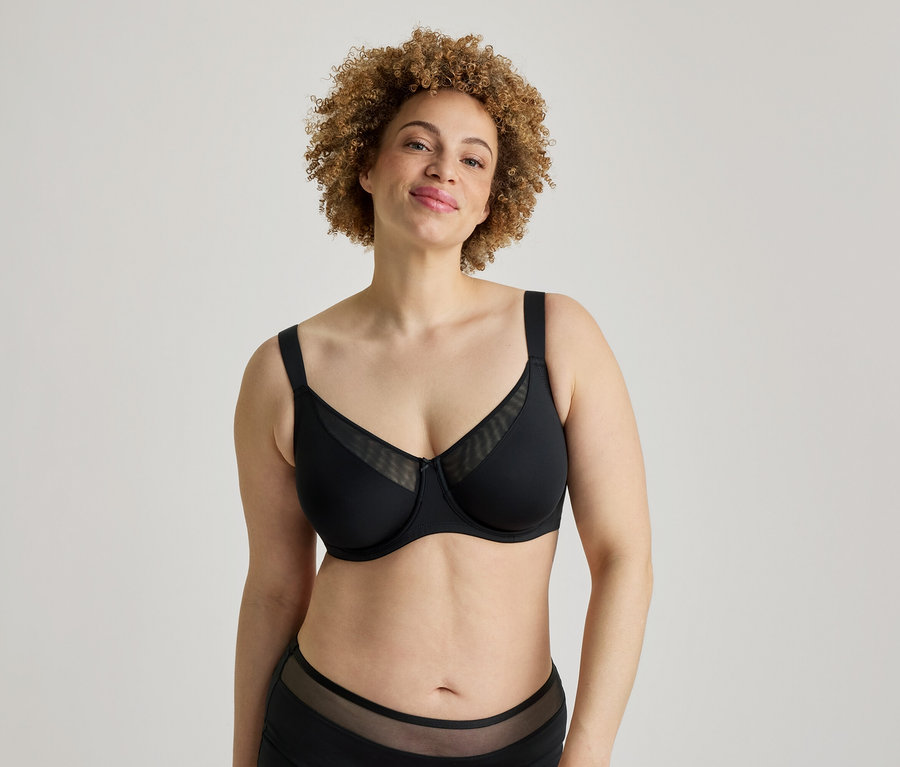 Eine lächelnde Frau mit lockigem Haar steht vor einem weißen Hintergrund und trägt einen schwarzen BH mit Netzeinsätzen und passender schwarzer Unterwäsche mit Netzdetails.