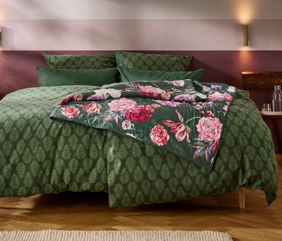 Ein Bett mit dunkelgrüner Bettwäsche mit Farnblattmuster. Eine gesteppte Blumendecke mit rosa und lila Blüten liegt über dem Fußende. Die Wand dahinter ist zweifarbig in Hellrosa und Dunkelburgund, mit zwei Wandleuchten. Links steht ein Holznachttisch mit einer rosa Blume in einer Vase, rechts ein Holzhocker mit einer Wasserkaraffe.