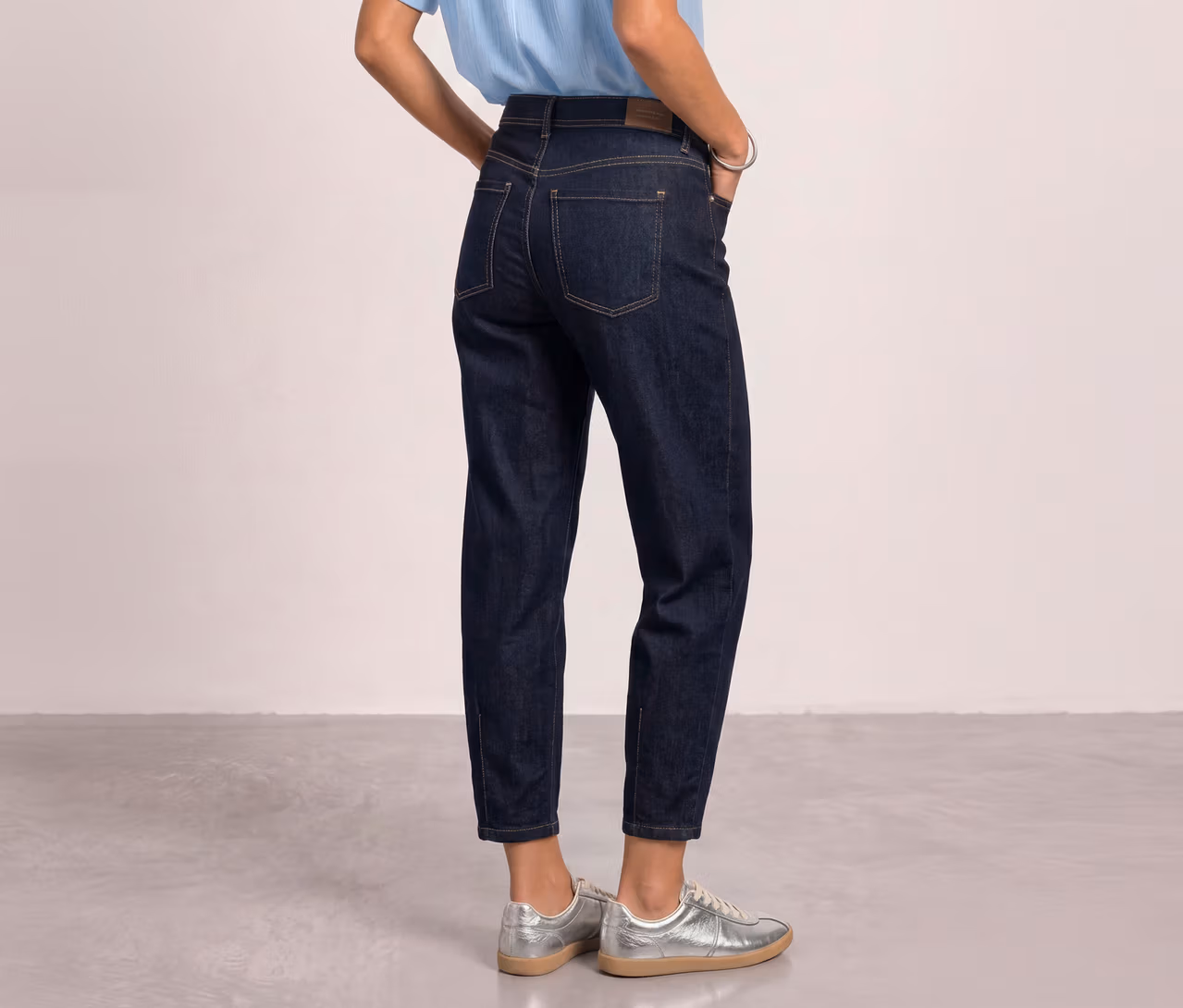 Person trägt dunkelblaue Street One Barrel Jeans, ein hellblaues Oberteil und silberne Sneaker, Hände in den Taschen.