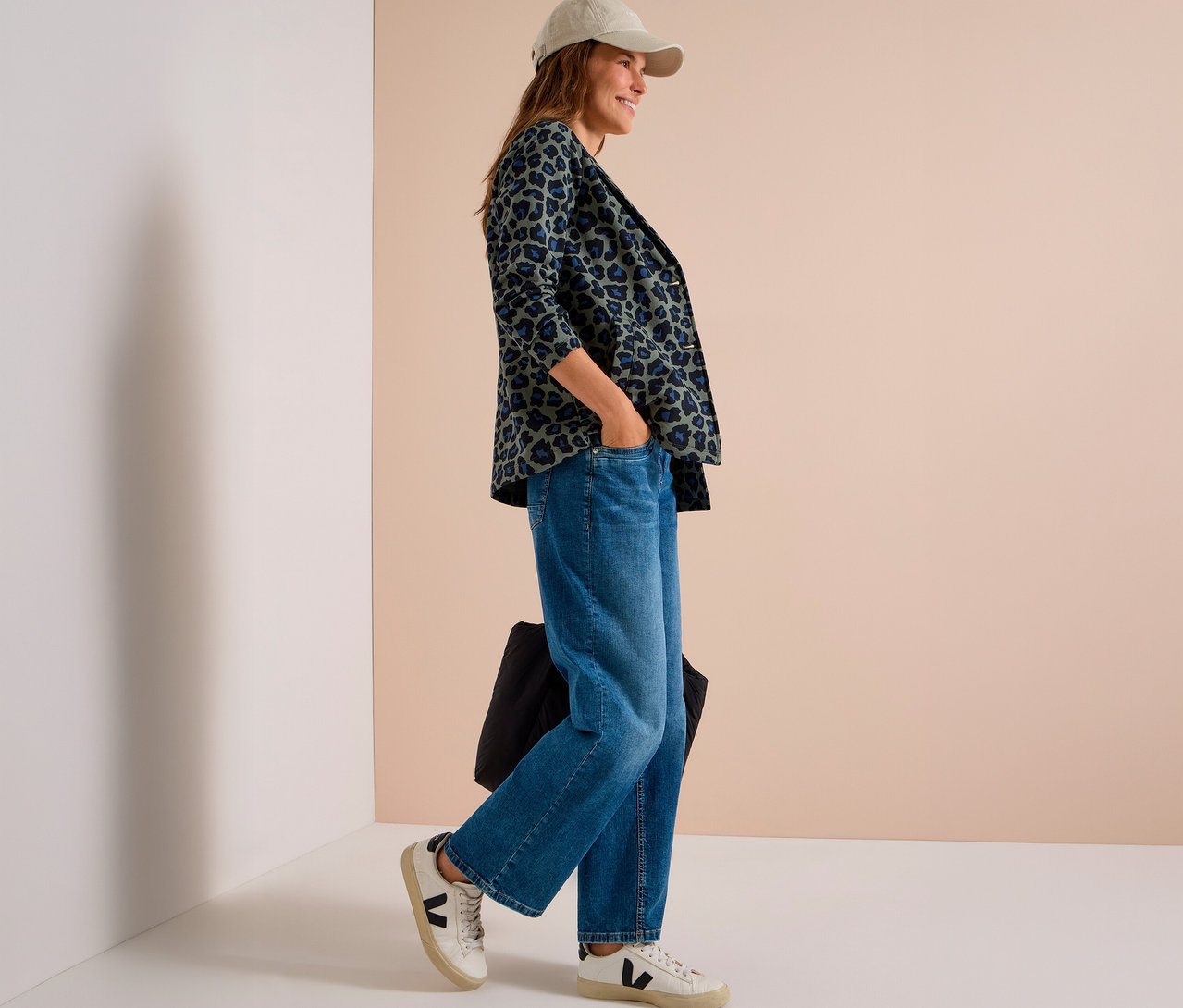 Frau trägt eine beigefarbene Kappe, einen grün-blauen CECIL Jerseyblazer mit Leopardenmuster, weite Jeans und weiße Sneaker, steht mit einer Hand in der Tasche und hält eine schwarze Tasche.