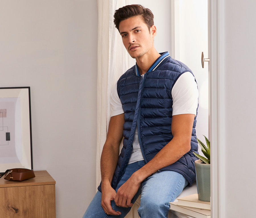 Mann sitzt mit blauer Steppweste auf Fensterbank.