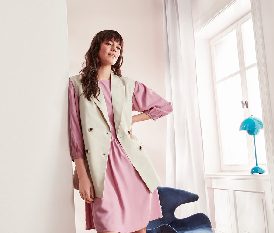 Frau lehnt an Wand und trägt rosa Kleid und Blazerweste.
