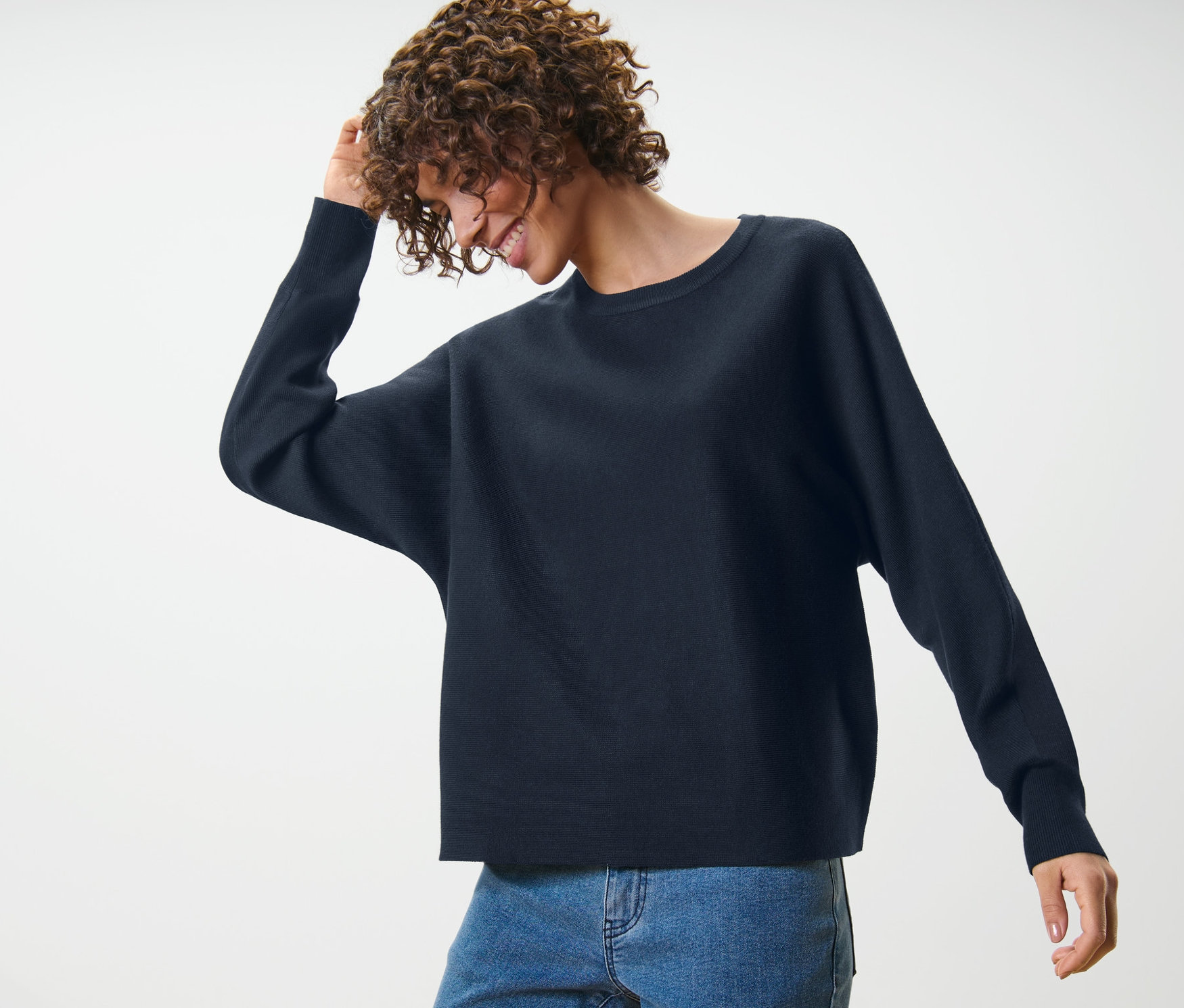 Frau mit lockigem Haar lächelt und berührt ihr Haar, sie trägt einen Feinstrickpullover mit Fledermausarm und blaue Jeans.