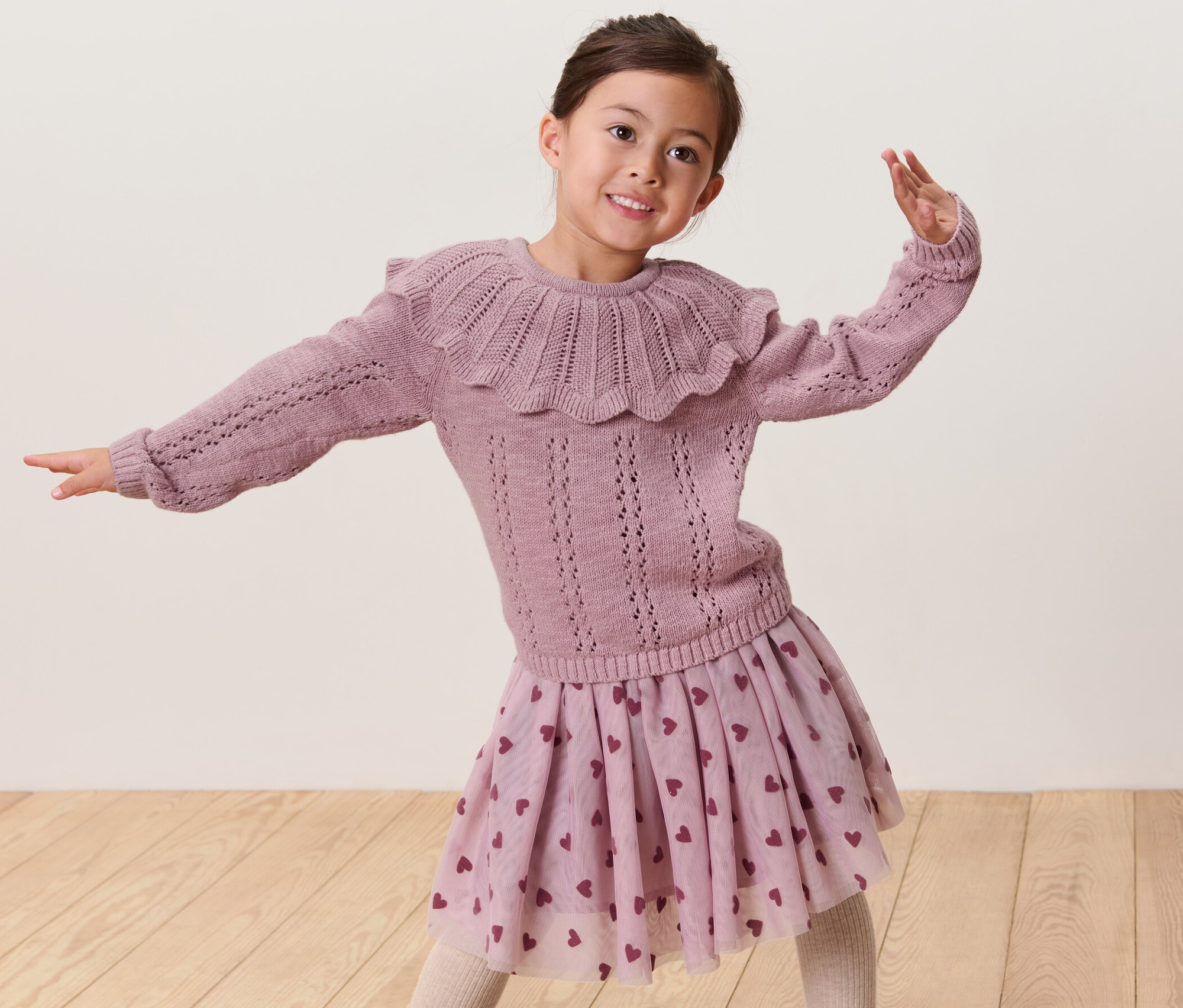 Lächelndes Mädchen posiert in einem Kinder-Strickpullover mit Kragen und einem Rock mit Herzchenmuster.