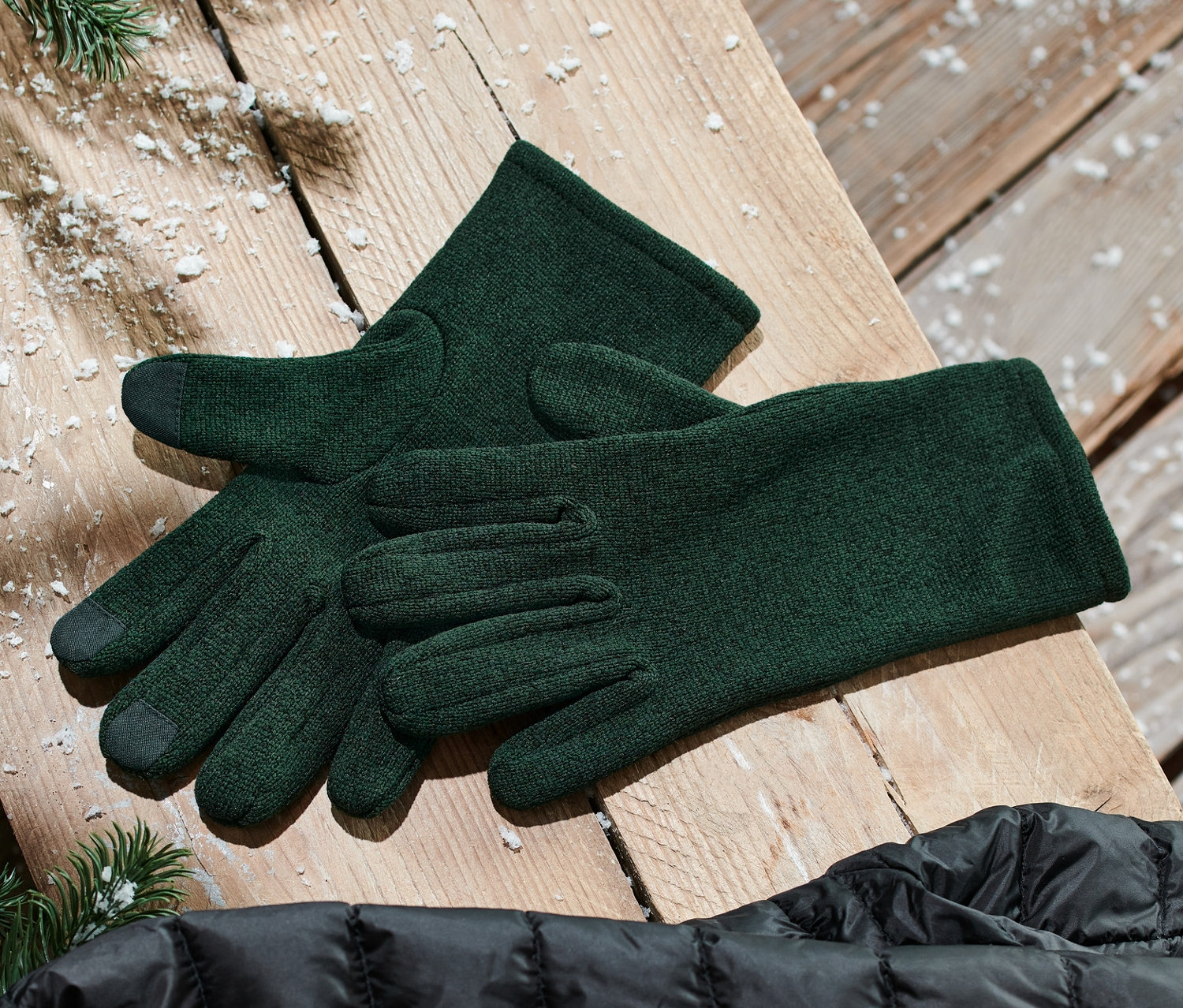 Grüne Strickfleece-Handschuhe liegen neben einer schwarzen Daunenjacke auf einer Holzfläche.