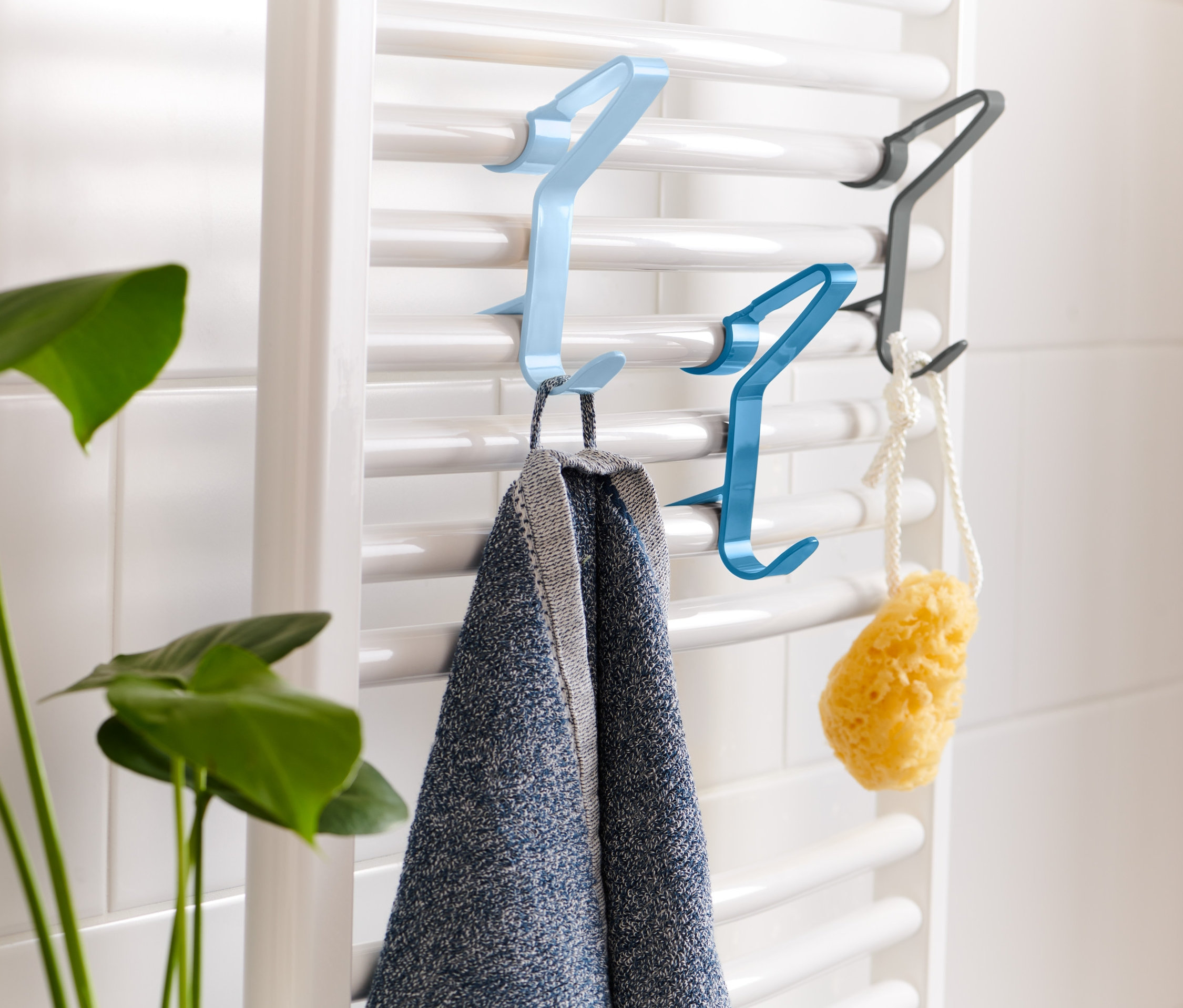 An einer Heizung hängen 3 Clip-Haken: hellblau, blau und schwarz. An dem hellblauen Haken hängt ein Handtuch, an dem blauen ein Schwamm.