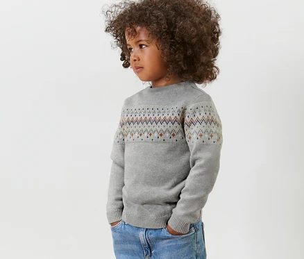 Seitenansicht eines Kindes mit lockigem Haar, das einen grauen Kinder-Strickpullover und Jeans trägt.