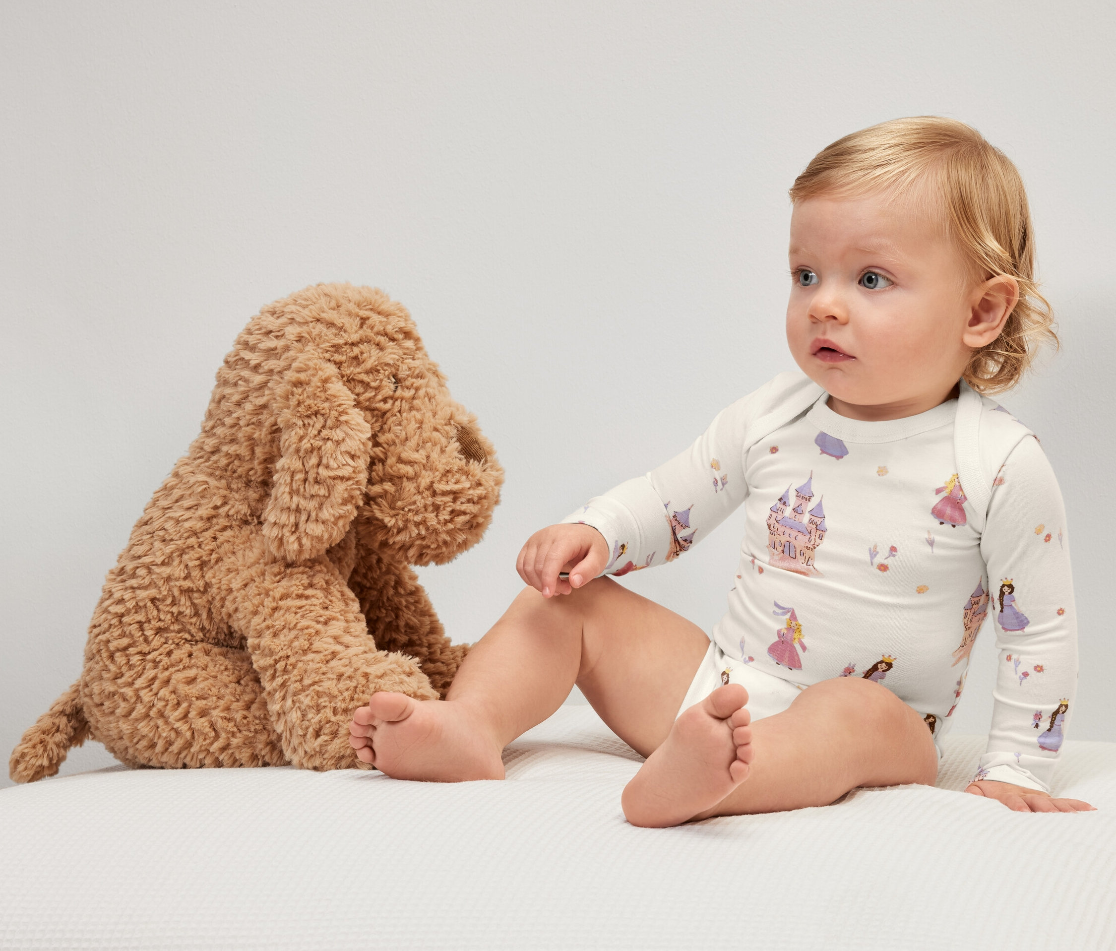 Baby sitzt neben Plüschhund und trägt einen mitwachsenden Body mit Prinzessinnenmotiv.