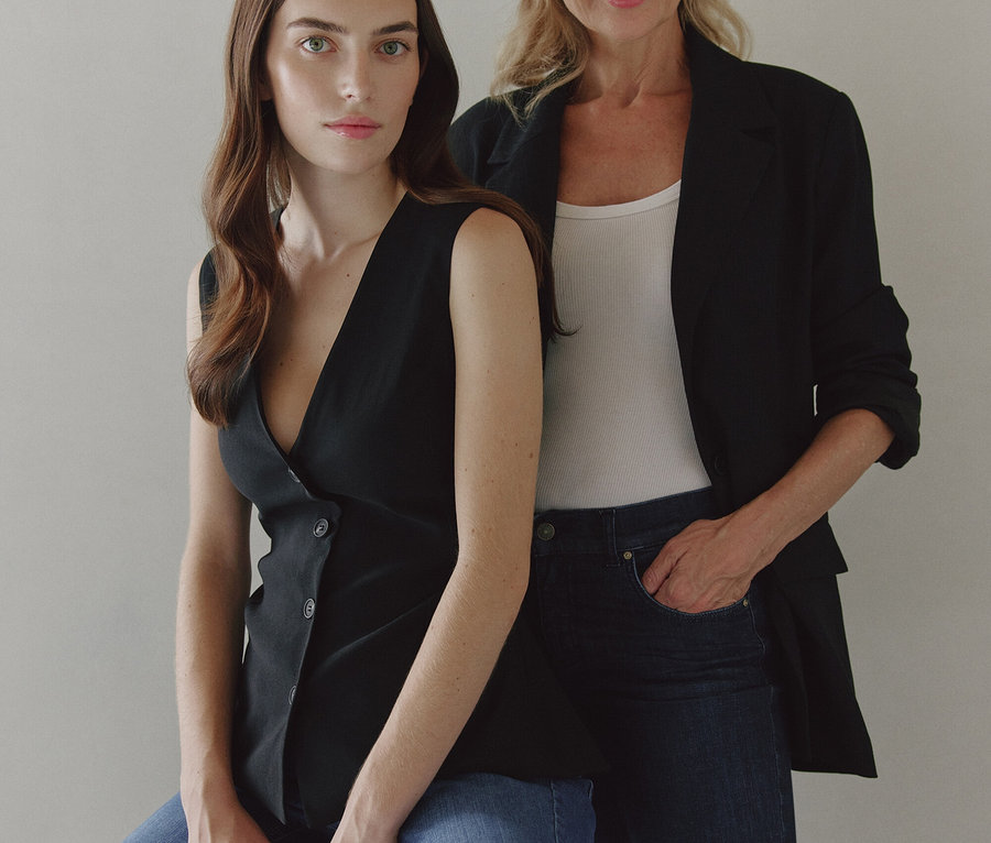 Zwei Frauen posieren. Eine sitzt auf einem Hocker in einem schwarzen Top und Jeans, die andere steht dahinter in einem schwarzen Blazer, einem weißen Tanktop und dunklen Jeans.