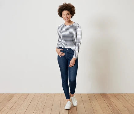 Frau mit lockigem Haar posiert in einem gestreiften Langarmshirt, dunkelblauen Jeans und weißen Turnschuhen.