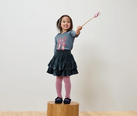 Ein lächelndes Mädchen hält einen Zauberstab und trägt ein blaues T-Shirt mit Ballettschuhen, einen grünen Rock und eine rosa Strumpfhose.