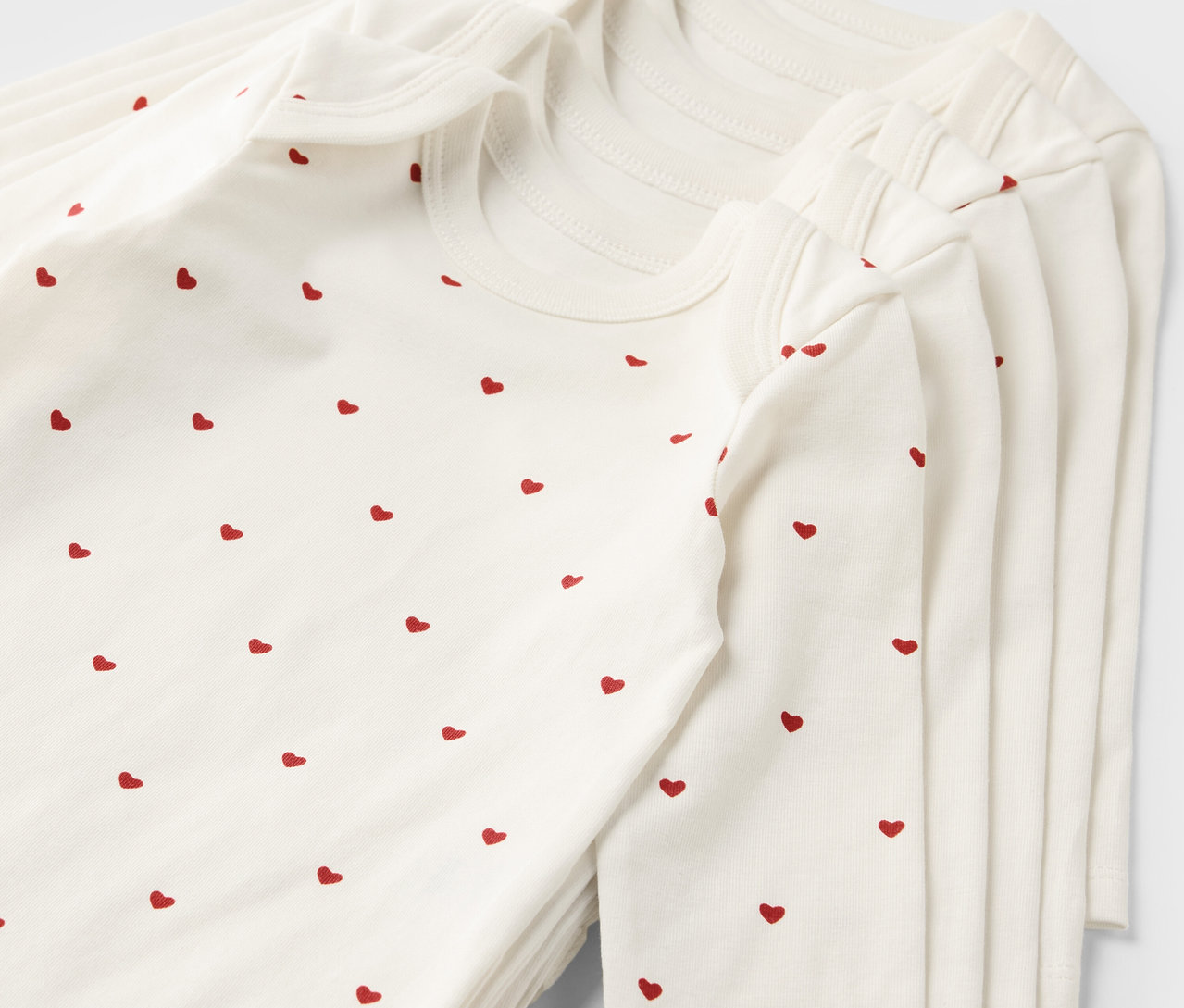Stapel aus fünf weißen Baby-Bodys mit Mitwachsfunktion und rotem Herzmuster.