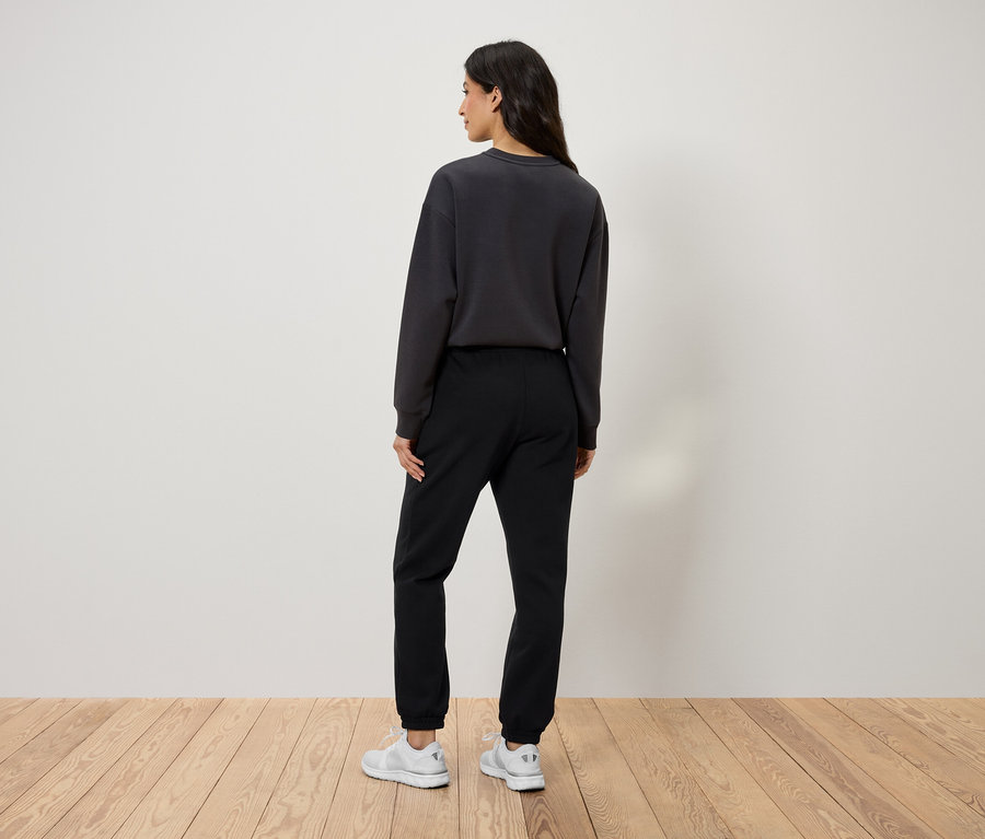 Rückansicht einer Frau in schwarzer Trainingshose, grauem Sweatshirt und weißen Schuhen.