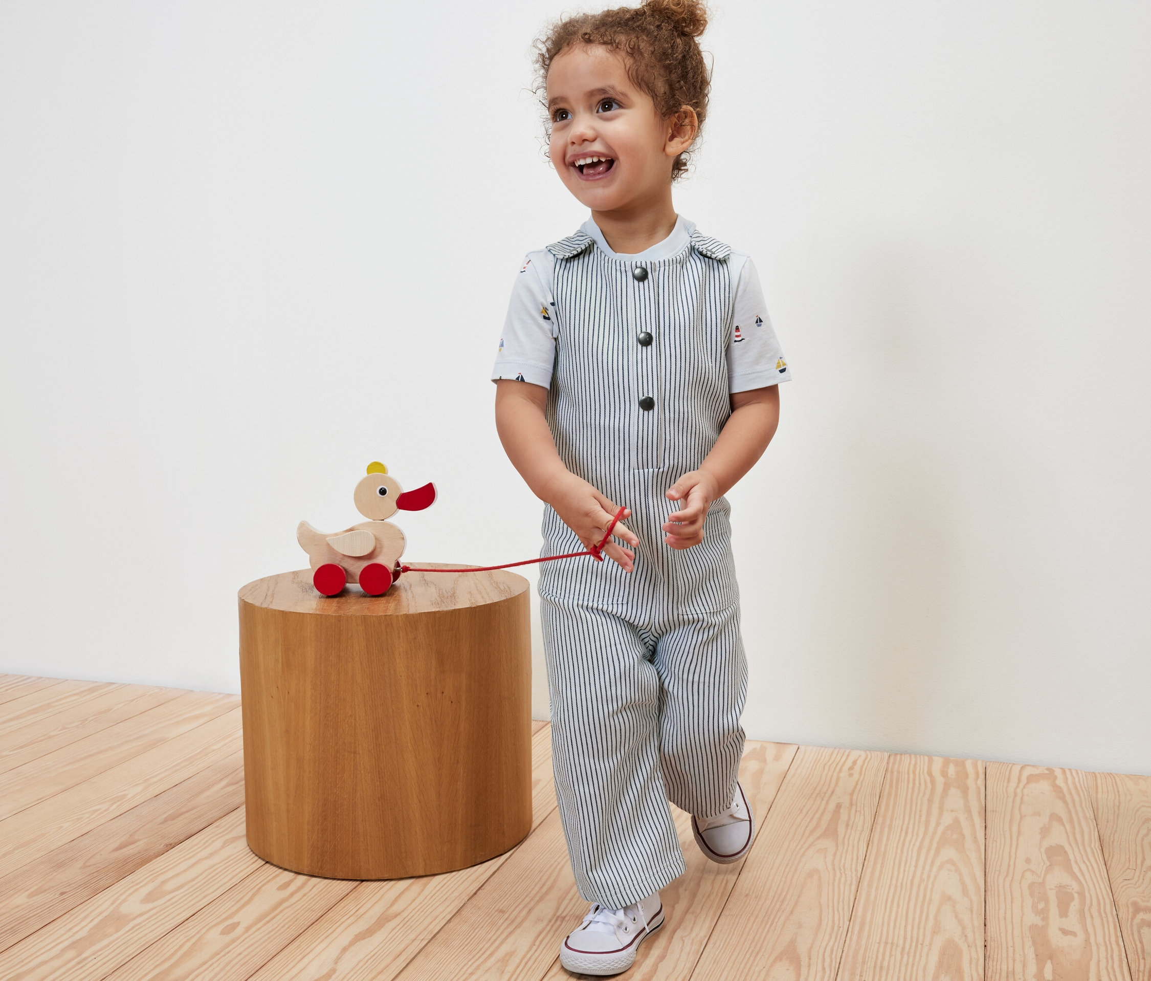 Lächelndes Kind zieht ein hölzernes Entenspielzeug auf Rädern und trägt eine Kinder-Web-Latzhose.